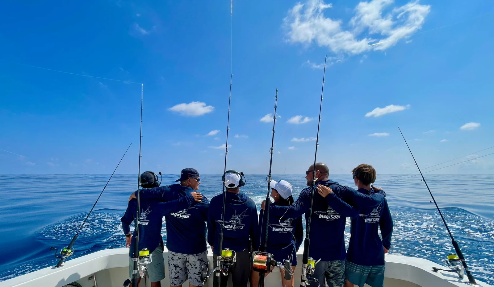 A crew in matching Vuelto Loco shirts standing at the bow of the charter boat with six fishing rods deployed under a blue sky.