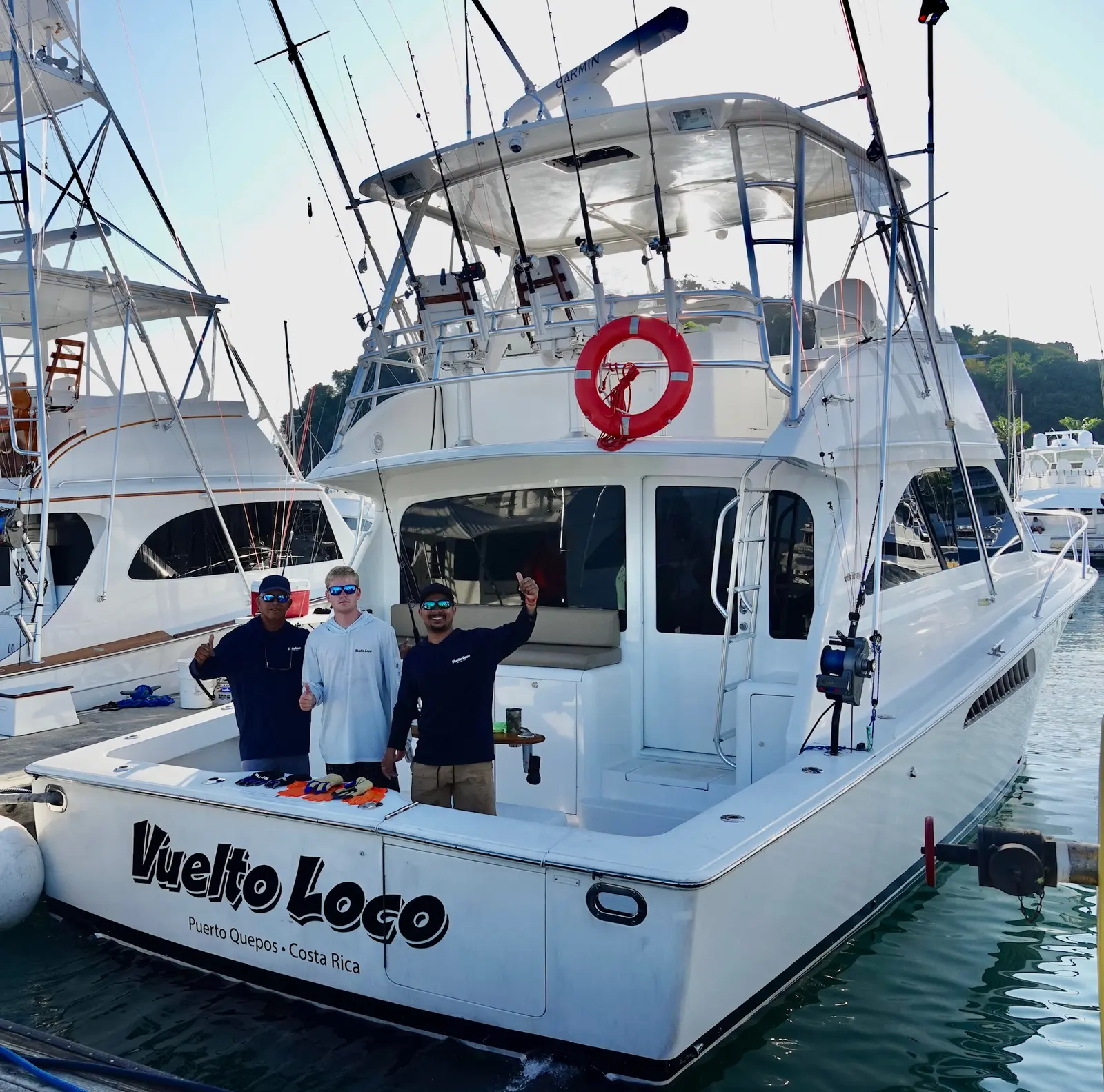 The sport-fishing boat Vuelto Loco docked at Marina Pez Vela in Quepos, seen from behind, with three people in the cockpit giving thumbs up and fishing gloves laid out on the transom.